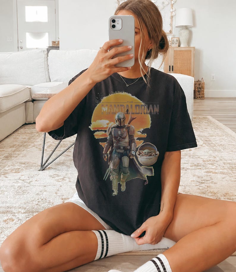K&ouml;nnte beinhalten: Ein schwarzes Kurzarm-T-Shirt mit einem Grafikdruck des Mandalorianers und Grogu in einer schwebenden Kapsel vor einem gelben Sonnenhintergrund. Der Text "THE MANDALORIAN" ist in einer abgenutzten Schrift &uuml;ber der Illustration gedruckt.