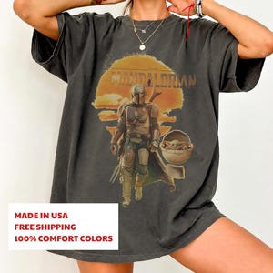 K&ouml;nnte beinhalten: Ein anthrazitfarbenes Kurzarm-T-Shirt mit einem Grafikdruck des Mandalorianers und Grogu in einer schwebenden Kapsel vor einer gro&szlig;en orangefarbenen Sonne. Der Text THE MANDALORIAN ist in einer verwaschenen Vintage-Schrift dar&uuml;ber gedruckt.