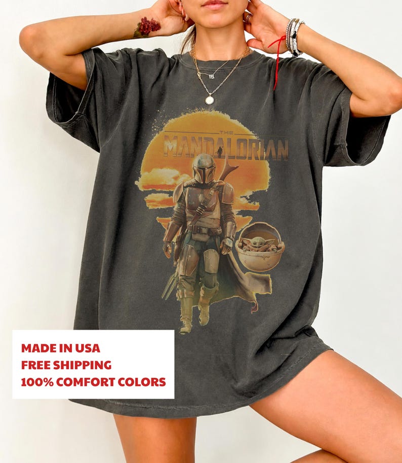 K&ouml;nnte beinhalten: Ein anthrazitfarbenes Kurzarm-T-Shirt mit einem Grafikdruck des Mandalorianers und Grogu in einer schwebenden Kapsel vor einer gro&szlig;en orangefarbenen Sonne. Der Text THE MANDALORIAN ist in einer verwaschenen Vintage-Schrift dar&uuml;ber gedruckt.