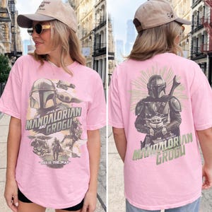 Puede incluir: Una camiseta rosa claro con gr&aacute;ficos de Star Wars. El frente muestra a The Mandalorian y Grogu con el texto "The Mandalorian and Grogu" y "This is the way". La parte posterior muestra una gran ilustraci&oacute;n de The Mandalorian sosteniendo a Grogu.