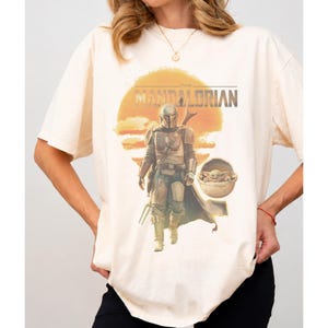 K&ouml;nnte beinhalten: Ein cremefarbenes Kurzarm-T-Shirt mit einem Grafikdruck des Mandalorianers und Grogu in einer schwebenden Kapsel vor einer gro&szlig;en orangefarbenen Sonne. Der Text THE MANDALORIAN ist in dunkelbraunen Buchstaben &uuml;ber der zentralen Figur gedruckt.