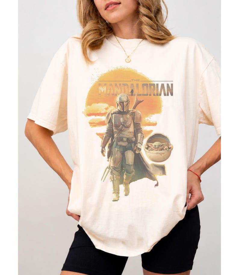 K&ouml;nnte beinhalten: Ein cremefarbenes Kurzarm-T-Shirt mit einem Grafikdruck des Mandalorianers und Grogu in einer schwebenden Kapsel vor einer gro&szlig;en orangefarbenen Sonne. Der Text THE MANDALORIAN ist in dunkelbraunen Buchstaben &uuml;ber der zentralen Figur gedruckt.