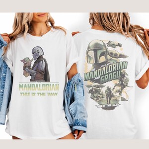 May include: A white t-shirt featuring Star Wars graphics. The front displays The Mandalorian holding Grogu with the text "The Mandalorian" and "This is the way". The back shows a collage of characters and vehicles with "The Mandalorian and Grogu" text.