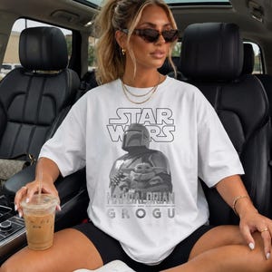 Może przedstawiać: Biały t-shirt z kr&oacute;tkim rękawem z czarno-białym nadrukiem graficznym przedstawiającym Mandalorianina i Grogu. Napis STAR WARS jest wydrukowany na czarno nad postaciami, a MANDALORIAN AND GROGU na szaro pod ilustracją.