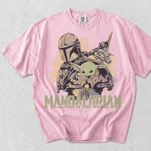 May include: A light pink short-sleeve t-shirt featuring a graphic print of The Mandalorian and Grogu. The design includes a spaceship, the text "MANDALORIAN" in large green letters, and the phrase "THIS IS THE WAY" printed underneath in smaller text.