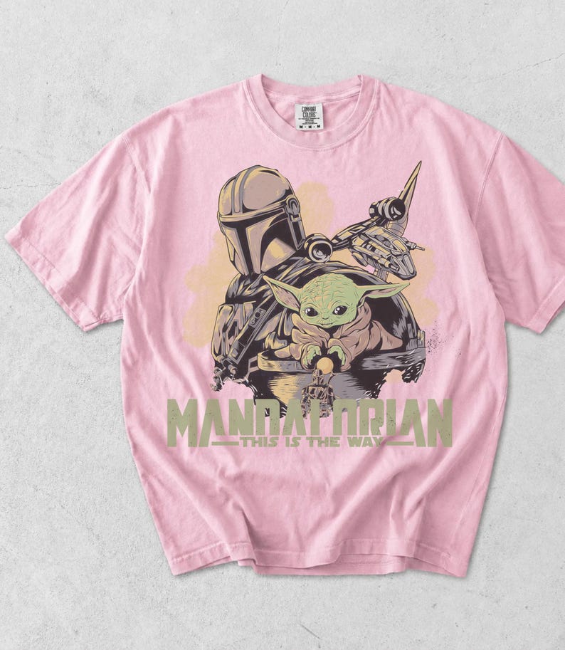 May include: A light pink short-sleeve t-shirt featuring a graphic print of The Mandalorian and Grogu. The design includes a spaceship, the text "MANDALORIAN" in large green letters, and the phrase "THIS IS THE WAY" printed underneath in smaller text.