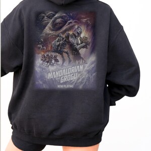 May include: A black hooded sweatshirt featuring a large graphic print on the back. The design depicts characters from The Mandalorian and Grogu, including a large creature eye, a helmeted figure, and small creatures. Text reads The Mandalorian and Grogu Now Playing.