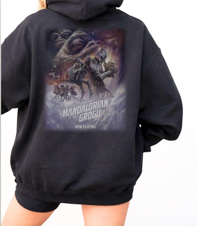 May include: A black hooded sweatshirt featuring a large graphic print on the back. The design depicts characters from The Mandalorian and Grogu, including a large creature eye, a helmeted figure, and small creatures. Text reads The Mandalorian and Grogu Now Playing.