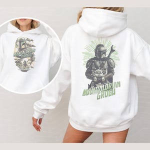 Puede incluir: Una sudadera con capucha blanca que presenta un estampado gr&aacute;fico de The Mandalorian y Grogu en la parte posterior y una versi&oacute;n m&aacute;s peque&ntilde;a en el frente. El dise&ntilde;o incluye a los personajes con armadura y t&uacute;nicas con texto verde.