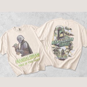 May include: Two off-white t-shirts featuring Star Wars graphics. The front shows The Mandalorian holding Grogu with the text "The Mandalorian This Is The Way". The back displays a larger collage of characters and the text "The Mandalorian and Grogu This Is The Way".
