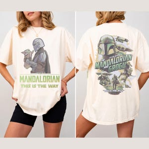 May include: An off-white t-shirt featuring a graphic of The Mandalorian and Grogu on the front with the text "The Mandalorian This Is The Way". The back displays a larger collage of the characters with the text "The Mandalorian and Grogu".