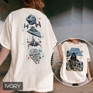 May include: An ivory t-shirt featuring a vertical graphic on the back with the Death Star, TIE fighters, a Star Destroyer, an X-wing, and the Millennium Falcon. The front displays a Star Wars logo above Darth Vader and various characters.