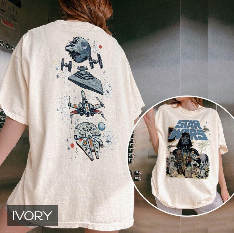 May include: An ivory t-shirt featuring a vertical graphic on the back with the Death Star, TIE fighters, a Star Destroyer, an X-wing, and the Millennium Falcon. The front displays a Star Wars logo above Darth Vader and various characters.