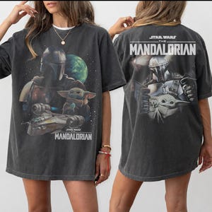 May include: A charcoal gray oversized t-shirt featuring a graphic print of The Mandalorian and Grogu. The front displays the characters with a spaceship and planet, while the back shows the characters with the text STAR WARS THE MANDALORIAN.
