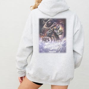 May include: A light gray hooded sweatshirt featuring a large rectangular graphic on the back. The print depicts characters from The Mandalorian and Grogu, with the text "THE MANDALORIAN AND GROGU" and "NOW PLAYING" printed in white below the artwork.
