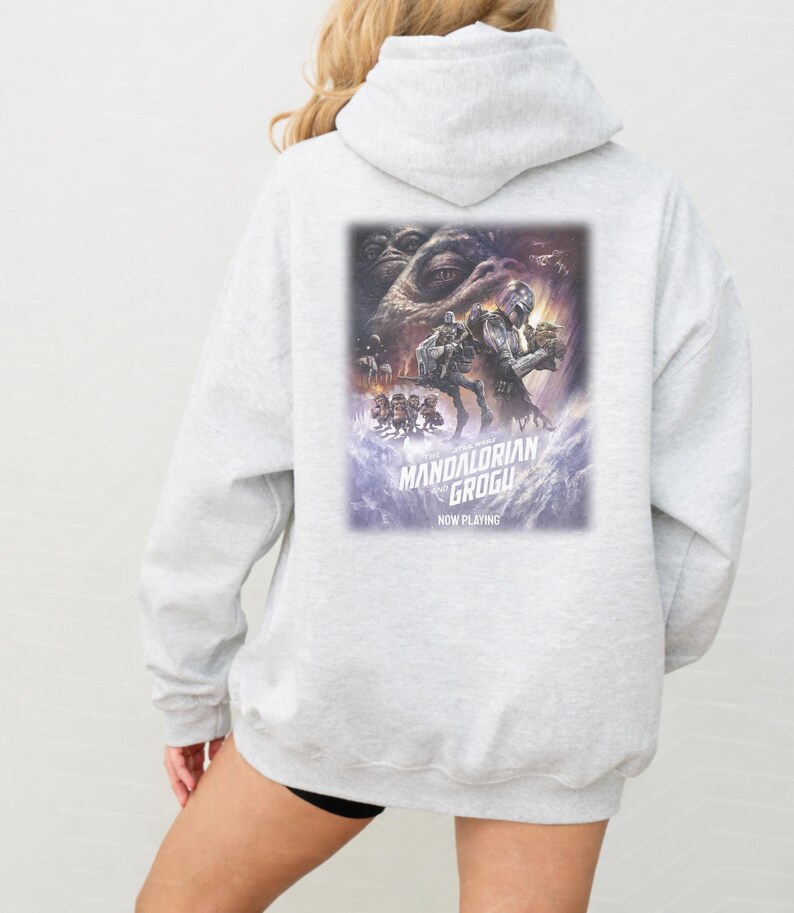 May include: A light gray hooded sweatshirt featuring a large rectangular graphic on the back. The print depicts characters from The Mandalorian and Grogu, with the text "THE MANDALORIAN AND GROGU" and "NOW PLAYING" printed in white below the artwork.