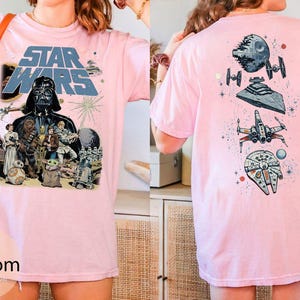 May include: A light pink t-shirt featuring a Star Wars graphic on the front with Darth Vader, Chewbacca, Princess Leia, and other characters. The back of the shirt displays a vertical arrangement of space vehicles including the Death Star and X-wing.