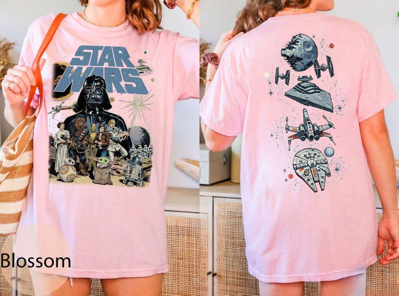 May include: A light pink t-shirt featuring a Star Wars graphic on the front with Darth Vader, Chewbacca, Princess Leia, and other characters. The back of the shirt displays a vertical arrangement of space vehicles including the Death Star and X-wing.
