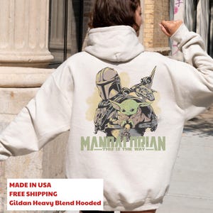 May include: A cream-colored hooded sweatshirt featuring a graphic on the back with a Mandalorian helmet, a small green creature, and a spacecraft. The text reads MANDALORIAN THIS IS THE WAY in green lettering. The hoodie has long sleeves and a hood.