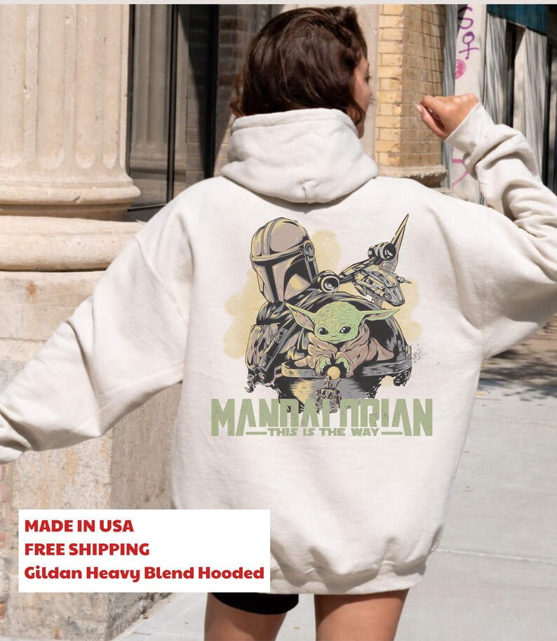 May include: A cream-colored hooded sweatshirt featuring a graphic on the back with a Mandalorian helmet, a small green creature, and a spacecraft. The text reads MANDALORIAN THIS IS THE WAY in green lettering. The hoodie has long sleeves and a hood.