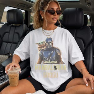May include: A white short-sleeved t-shirt featuring a graphic print of The Mandalorian and Grogu. The design includes the text Star Wars The Mandalorian and Grogu in yellow and black lettering. The shirt has a crew neckline and a relaxed fit.