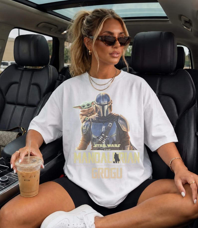May include: A white short-sleeved t-shirt featuring a graphic print of The Mandalorian and Grogu. The design includes the text Star Wars The Mandalorian and Grogu in yellow and black lettering. The shirt has a crew neckline and a relaxed fit.