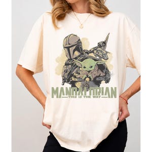 May include: A cream-colored short-sleeve t-shirt featuring a graphic print of a helmeted character and a small green creature. The text "MANDALORIAN" and "THIS IS THE WAY" is printed in green below the illustration, which also includes a spaceship.