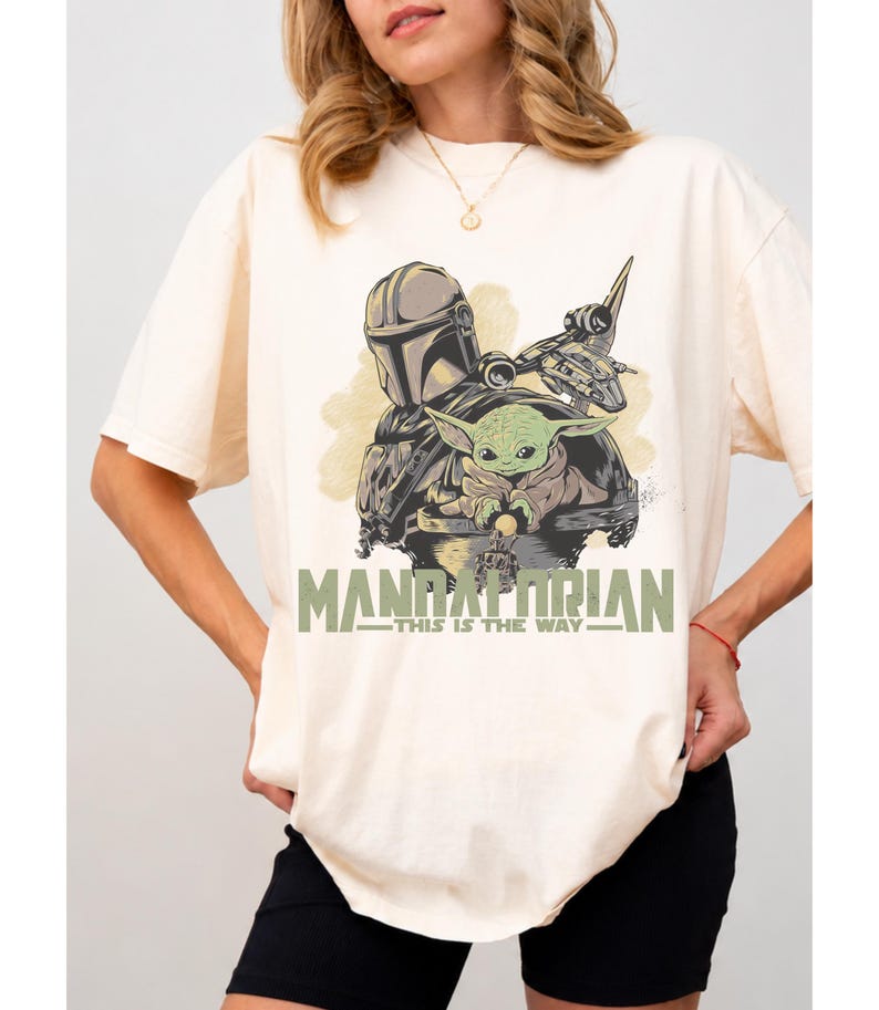 May include: A cream-colored short-sleeve t-shirt featuring a graphic print of a helmeted character and a small green creature. The text "MANDALORIAN" and "THIS IS THE WAY" is printed in green below the illustration, which also includes a spaceship.