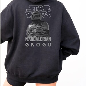 Może przedstawiać: Czarna bluza z kapturem z grafiką na plecach przedstawiającą napis STAR WARS u g&oacute;ry, ilustrację Mandalorianina trzymającego Grogu na środku oraz napis THE MANDALORIAN AND GROGU wydrukowany w kolorze szarym pod postaciami.