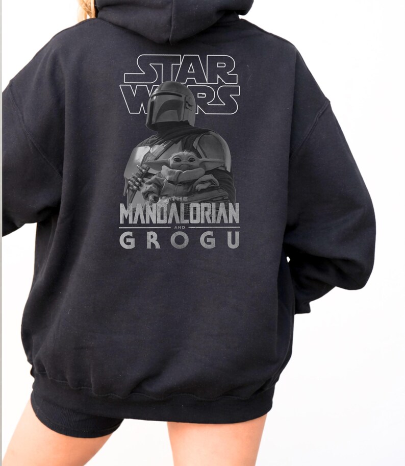 Może przedstawiać: Czarna bluza z kapturem z grafiką na plecach przedstawiającą napis STAR WARS u g&oacute;ry, ilustrację Mandalorianina trzymającego Grogu na środku oraz napis THE MANDALORIAN AND GROGU wydrukowany w kolorze szarym pod postaciami.