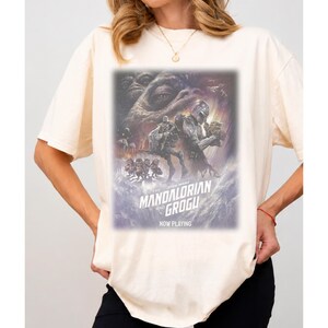 May include: A cream-colored short-sleeve t-shirt featuring a graphic print of The Mandalorian and Grogu. The design includes the text "The Mandalorian and Grogu" and "Now Playing" with a cinematic illustration of characters from the Star Wars universe.