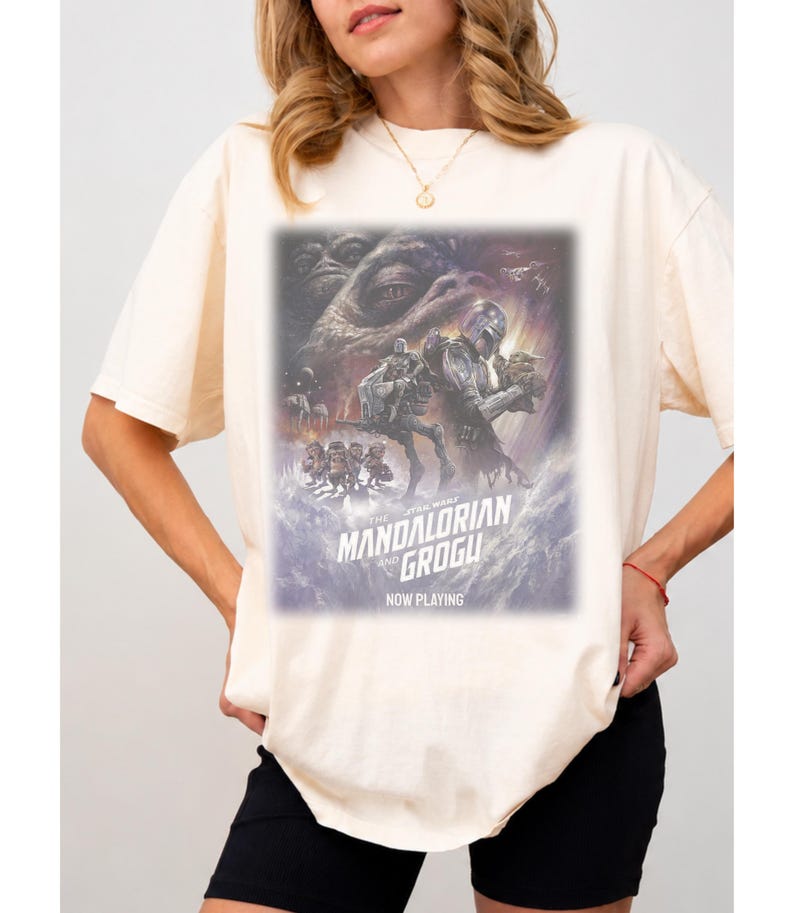 May include: A cream-colored short-sleeve t-shirt featuring a graphic print of The Mandalorian and Grogu. The design includes the text "The Mandalorian and Grogu" and "Now Playing" with a cinematic illustration of characters from the Star Wars universe.