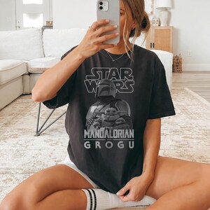 Może przedstawiać: Czarny t-shirt z kr&oacute;tkim rękawem z białym nadrukiem graficznym logo Star Wars nad obrazem Mandalorianina trzymającego Grogu. Tekst pod obrazem brzmi The Mandalorian and Grogu pogrubioną, białą czcionką bezszeryfową.