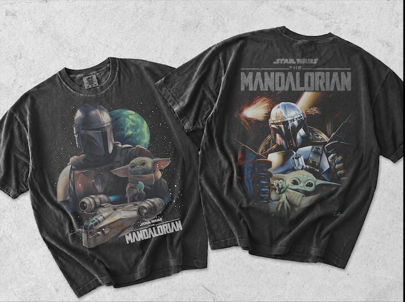 May include: Two black short-sleeve t-shirts featuring graphic prints from Star Wars The Mandalorian. The designs display the Mandalorian character and Grogu in space settings with the text Star Wars The Mandalorian printed on the fabric.