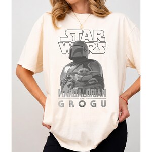 Może przedstawiać: Kremowy t-shirt z kr&oacute;tkim rękawem z czarno-białą grafiką przedstawiającą Mandalorianina trzymającego Grogu. Na g&oacute;rze znajduje się napis STAR WARS, a pod ilustracją postaci widnieje pogrubiony, postarzany napis MANDALORIAN AND GROGU.