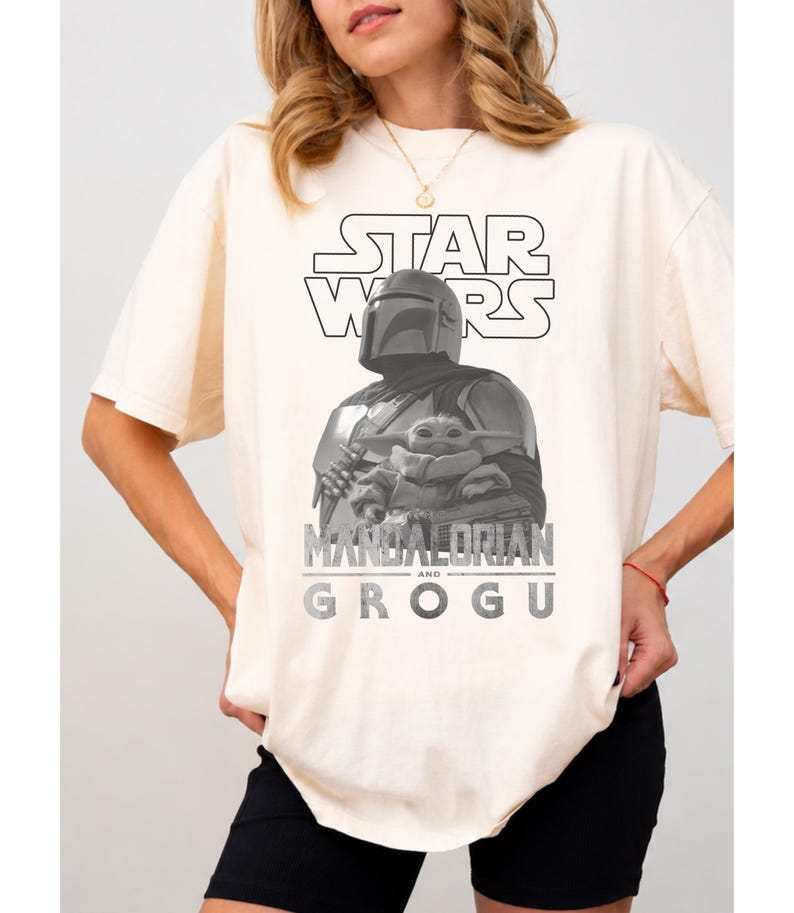 Może przedstawiać: Kremowy t-shirt z kr&oacute;tkim rękawem z czarno-białą grafiką przedstawiającą Mandalorianina trzymającego Grogu. Na g&oacute;rze znajduje się napis STAR WARS, a pod ilustracją postaci widnieje pogrubiony, postarzany napis MANDALORIAN AND GROGU.