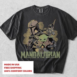 May include: A charcoal grey short-sleeve t-shirt featuring a graphic print of a helmeted warrior and a small green creature in a floating pod. The text "MANDALORIAN" and "THIS IS THE WAY" is printed in olive green below the central character illustration.