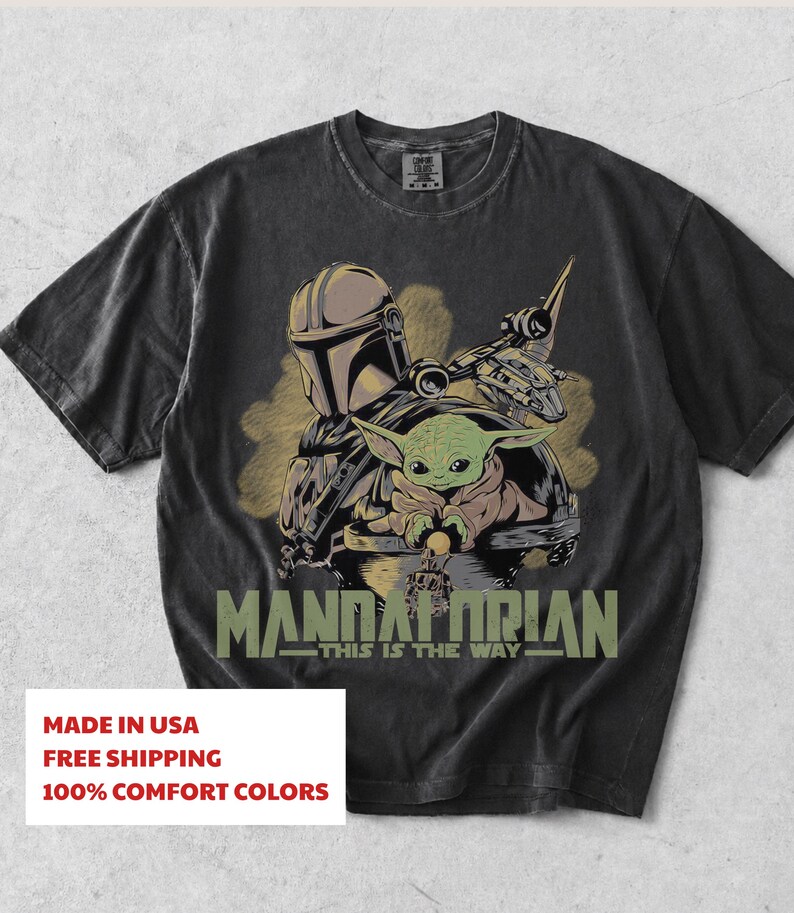 May include: A charcoal grey short-sleeve t-shirt featuring a graphic print of a helmeted warrior and a small green creature in a floating pod. The text "MANDALORIAN" and "THIS IS THE WAY" is printed in olive green below the central character illustration.