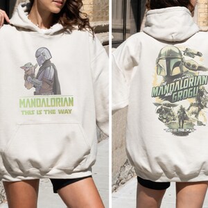 May include: An off-white hooded sweatshirt featuring a graphic of The Mandalorian and Grogu on the front with the text The Mandalorian This Is The Way. The back displays a larger, detailed collage of the characters and the text This Is The Way.