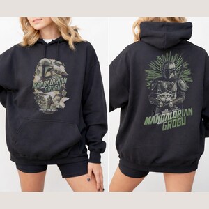 Puede incluir: Una sudadera con capucha negra con un estampado gr&aacute;fico de The Mandalorian y Grogu. El frente muestra un collage con el texto "The Mandalorian and Grogu" y "This is the way". La parte posterior muestra una gran ilustraci&oacute;n de los personajes.