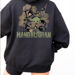 May include: A black hooded sweatshirt featuring a graphic on the back with a Mandalorian character, a small green creature, and a spacecraft. The text "MANDALORIAN" and "THIS IS THE WAY" is printed in olive green below the illustrated characters.