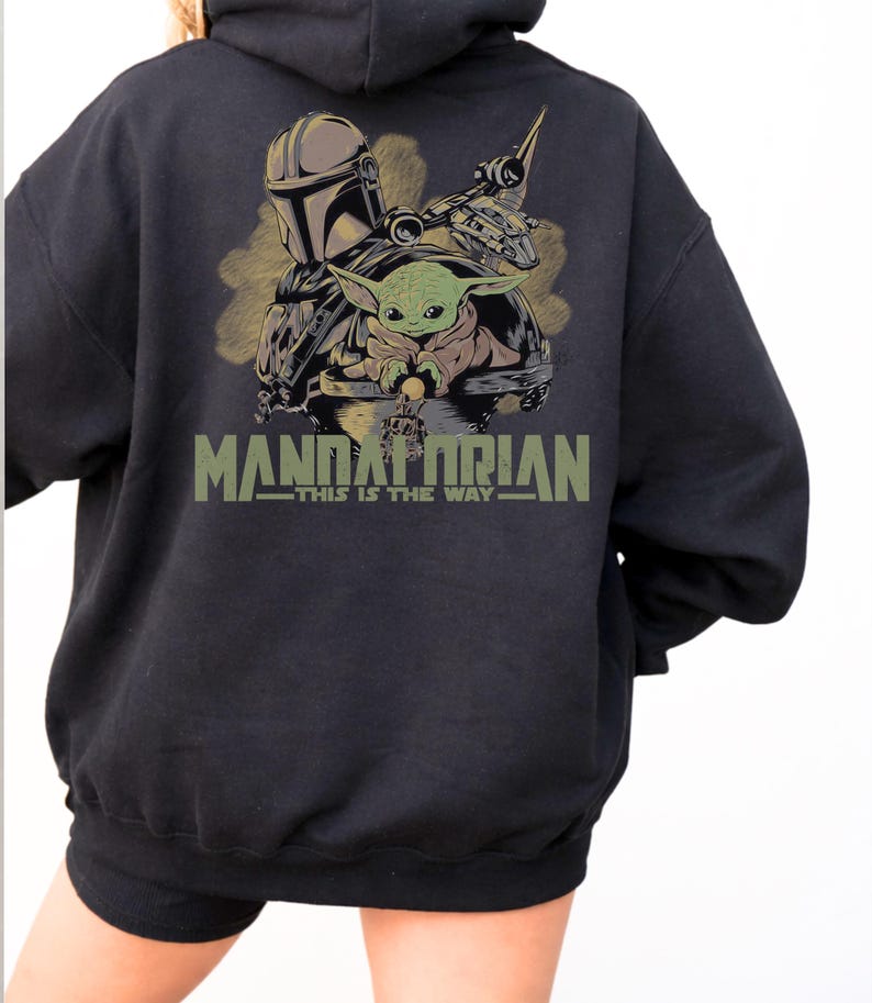 May include: A black hooded sweatshirt featuring a graphic on the back with a Mandalorian character, a small green creature, and a spacecraft. The text "MANDALORIAN" and "THIS IS THE WAY" is printed in olive green below the illustrated characters.