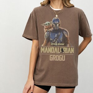 May include: A brown short-sleeve t-shirt featuring a graphic print of The Mandalorian and Grogu. The text reads Star Wars The Mandalorian and Grogu in a tan font. The shirt has a crew neckline and a relaxed fit, showcasing the characters in their armor.