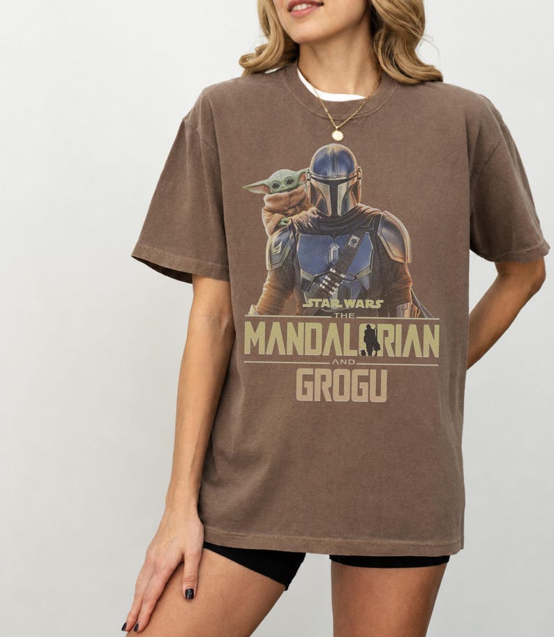 May include: A brown short-sleeve t-shirt featuring a graphic print of The Mandalorian and Grogu. The text reads Star Wars The Mandalorian and Grogu in a tan font. The shirt has a crew neckline and a relaxed fit, showcasing the characters in their armor.