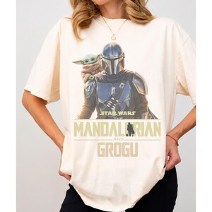 May include: A cream-colored short-sleeve t-shirt featuring a graphic print of The Mandalorian and Grogu. The text reads Star Wars The Mandalorian and Grogu in a bold, tan-colored font. The design shows the armored character with the small creature.