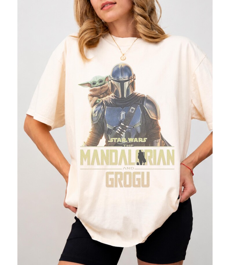 May include: A cream-colored short-sleeve t-shirt featuring a graphic print of The Mandalorian and Grogu. The text reads Star Wars The Mandalorian and Grogu in a bold, tan-colored font. The design shows the armored character with the small creature.