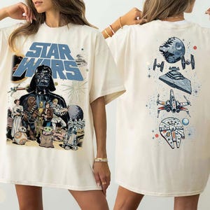 May include: An ivory t-shirt featuring a large Star Wars graphic on the front with Darth Vader, Luke Skywalker, Princess Leia, Chewbacca, and other characters. The back displays a vertical arrangement of space vehicles including the Death Star.