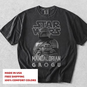 Może przedstawiać: Ciemnoszary t-shirt z kr&oacute;tkim rękawem z białym nadrukiem graficznym przedstawiającym Mandalorianina i Grogu. Nad postaciami znajduje się napis STAR WARS, a poniżej THE MANDALORIAN AND GROGU. Koszulka posiada okrągły dekolt.