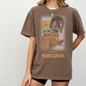 May include: A brown short-sleeve t-shirt featuring a graphic print of The Mandalorian and Grogu. The design includes a yellow sun, a spacecraft, and the text STAR WARS THE MANDALORIAN in a tan font. The shirt has a crew neckline and a relaxed fit.