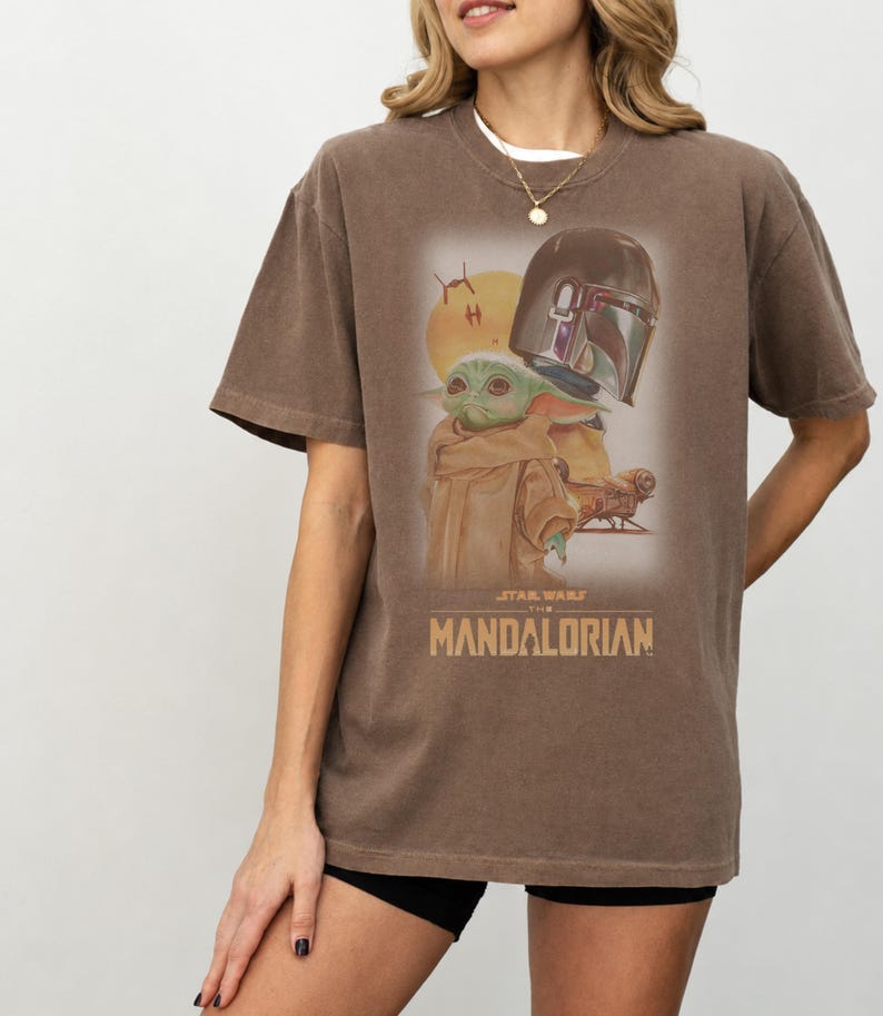 May include: A brown short-sleeve t-shirt featuring a graphic print of The Mandalorian and Grogu. The design includes a yellow sun, a spacecraft, and the text STAR WARS THE MANDALORIAN in a tan font. The shirt has a crew neckline and a relaxed fit.